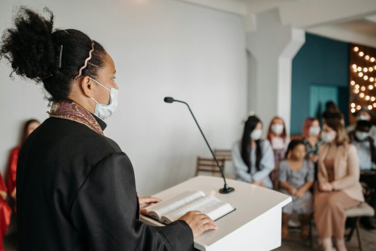 Female pastor delivering a sermon to a masked congregation indoors.